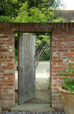 an open wooden door in a brick wall next to a potted plant and tree