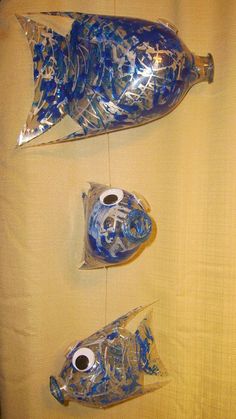 three blue glass fish hanging from strings on a yellow wall with white fabric behind them