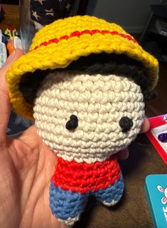 a small crocheted doll with a yellow hat and red shirt on it's head