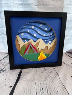 a picture frame with a campfire and mountains in the background on a wooden table