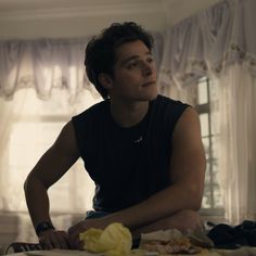 a young man sitting on top of a bed next to a white curtained window