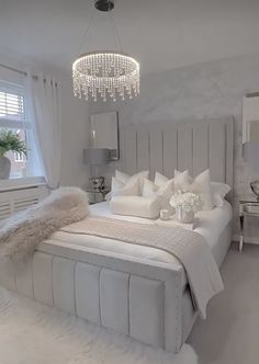 a white bedroom with a chandelier hanging from the ceiling and pillows on the bed