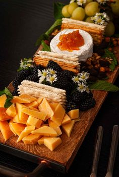 cheese, crackers and fruit are arranged on a cutting board with utensils