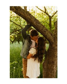 a man and woman kissing under a tree in the woods with green grass behind them