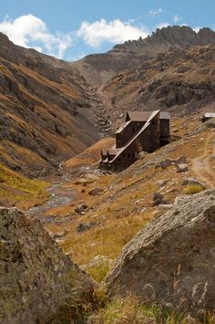 an old building in the middle of a mountain range with rocks and grass around it