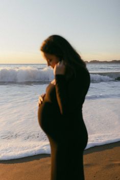 a pregnant woman is standing on the beach talking on her cell phone while looking at the ocean