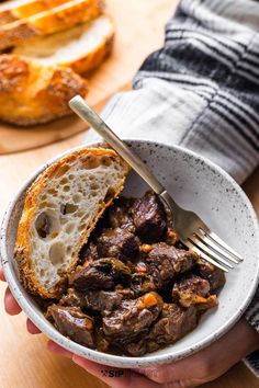 a person holding a white bowl filled with meat and bread on top of a wooden table