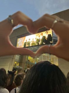 two hands making a heart shape in front of a building with people standing around it