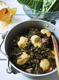 a pot filled with greens and dumplings on top of a white table next to a wooden spoon