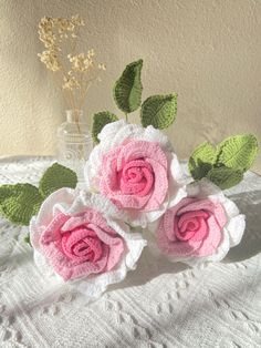 three crocheted roses are sitting on a white tablecloth next to a vase with flowers in it