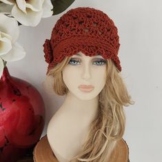 a mannequin head wearing a red crochet hat next to a vase with white flowers