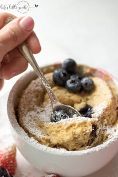 a hand holding a spoon over a bowl of blueberry cheesecake with powdered sugar