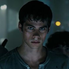 a young man with blue eyes stares at the camera in a dimly lit room as people look on