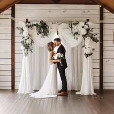 a bride and groom standing in front of a wedding arch with white flowers on it