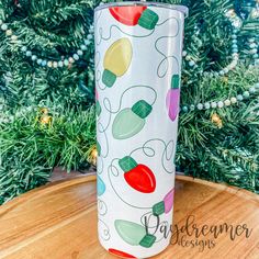 a white tumbler with christmas lights on it sitting in front of a pine tree