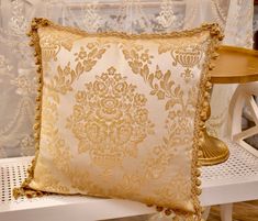 a gold pillow on a white bench in front of a window with lace curtain behind it