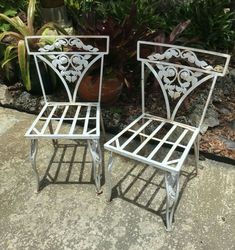 two white metal chairs sitting next to each other on a cement ground in front of plants