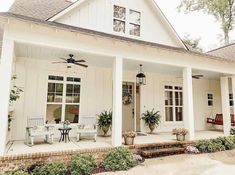 a white house with lots of windows and plants on the front porch, along with two chairs