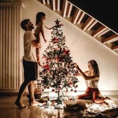 two people decorating a christmas tree in front of a stair case, while another person stands near the top