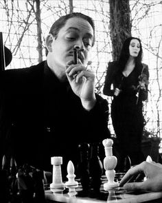 a man is playing chess with two women in the background and one woman standing behind him