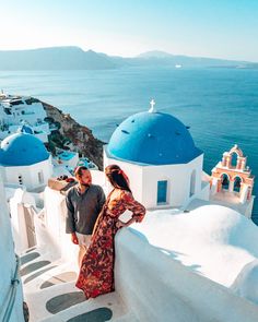 a man and woman standing on top of a white building next to the ocean with blue domes