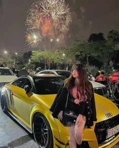 a woman sitting on the hood of a yellow sports car with fireworks in the background