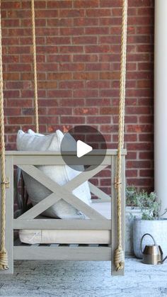 a porch swing made out of wood and rope with pillows on the bottom, sitting in front of a brick wall