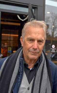 an older man with grey hair wearing a gray and black striped scarf in front of a building