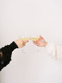 two people toasting with wine glasses in front of a white wall and one person is holding out their hand