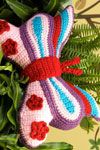 a crocheted butterfly ornament sitting on top of a green plant in front of a wall