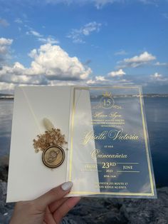 a person holding up a wedding card in front of the water with clouds and blue sky