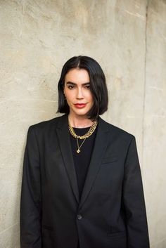 a woman wearing a black suit and gold necklace standing in front of a concrete wall