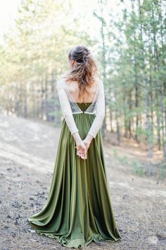 a woman in a long green dress standing on a dirt road with her back to the camera