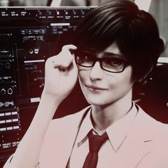 a woman wearing glasses and a tie in front of a computer screen with other electronic equipment behind her