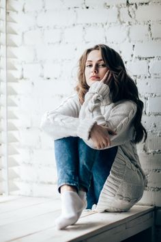 a woman sitting on top of a wooden bench in front of a brick wall with her legs crossed