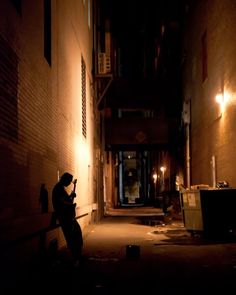 a person sitting on a bench in an alleyway at night with their cell phone up against the wall