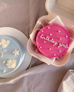 two birthday cakes in plastic containers on a table with pink and white flowers around them