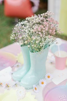 a table with flowers in rain boots and plates on the tablecloth, next to a pink vase filled with baby's breath