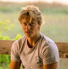 a young man sitting on top of a wooden bench next to a lush green field