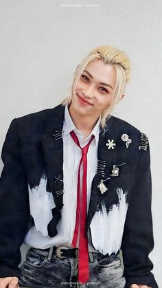 a man with blonde hair wearing a black jacket and red tie, sitting on a chair