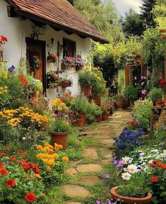 a garden filled with lots of flowers next to a white house on a dirt road