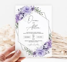 a person holding up a wedding card with purple flowers and greenery in the background