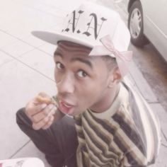 a young man wearing a hat and eating food on the side walk near a parked car