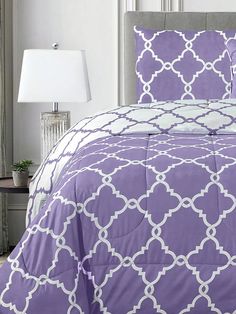 a bed with purple and white comforters in a room next to a lamp on a table