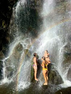 three women in bathing suits standing at the base of a waterfall with a rainbow behind them