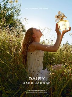 a woman sitting in tall grass holding up a perfume bottle