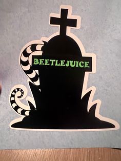 a hand holding up a sticker that says beetlejuice with a grave in the background