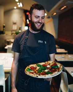 a man in an apron holding a pizza on a plate with basil and tomato sauce