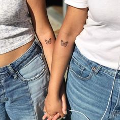 two women holding hands with butterfly tattoos on their arms and wristbands, both wearing blue jeans