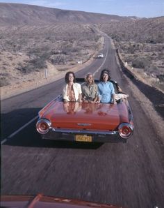 three people are sitting in the back of an old red convertible car on a desert road